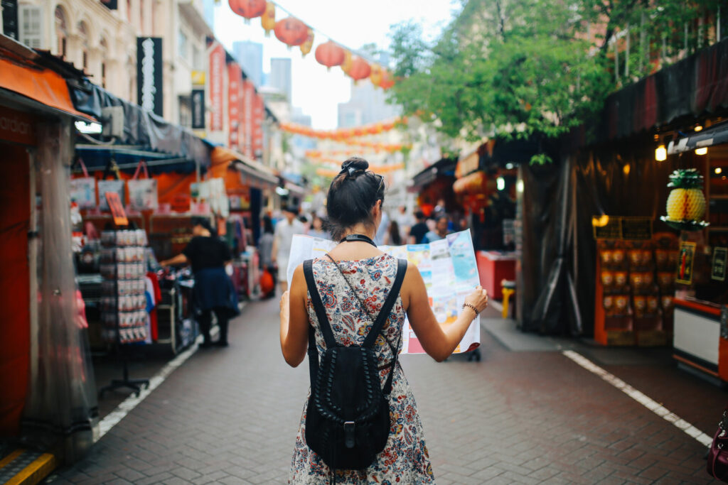 Rear view image of a young brunette woman. She is enjoying the walk and exploring the city, wearing a casual but fashionable dress, sightseeing and shopping on the Singapore street market. She is holding a large city map, checking out where to go next.